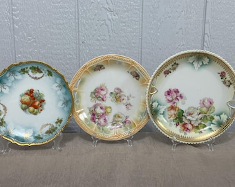 Könnte beinhalten: Drei antike Porzellanteller mit goldenen Rändern und Blumendesigns. Die Teller zeigen verschiedene Blumenmuster, darunter Rosen und andere Blumen. Die Teller werden auf weißen Ständern ausgestellt.