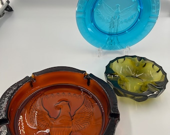 May include: Three glass ashtrays with an eagle design. The ashtrays are in shades of blue, brown, and green.