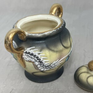 May include: A small, cream-colored ceramic sugar bowl with a gold lid and handles. The bowl is decorated with a black and white dragon design and blue accents.