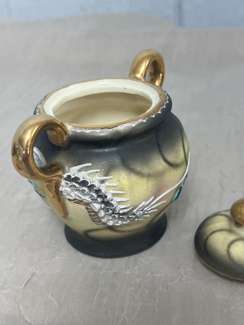 May include: A small, cream-colored ceramic sugar bowl with a gold lid and handles. The bowl is decorated with a black and white dragon design and blue accents.