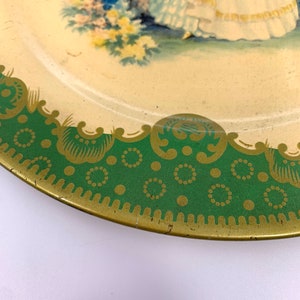 May include: A vintage tin plate with a green and gold border. The center of the plate features a painted image of a woman in a white dress standing in a garden with flowers.