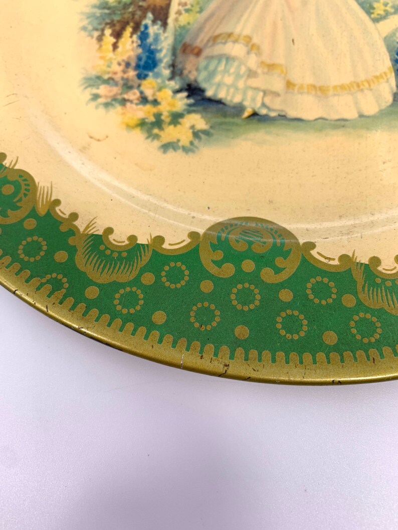 May include: A vintage tin plate with a green and gold border. The center of the plate features a painted image of a woman in a white dress standing in a garden with flowers.