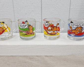 May include: Set of four glass mugs featuring Garfield the cat. Each mug has a different cartoon image of Garfield with a humorous quote. The mugs are clear glass with a white rim and handle.