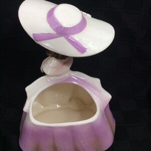 May include: A ceramic planter shaped like a woman wearing a white hat with a purple ribbon. The planter has a purple and white glaze and is designed to hold small plants or flowers.