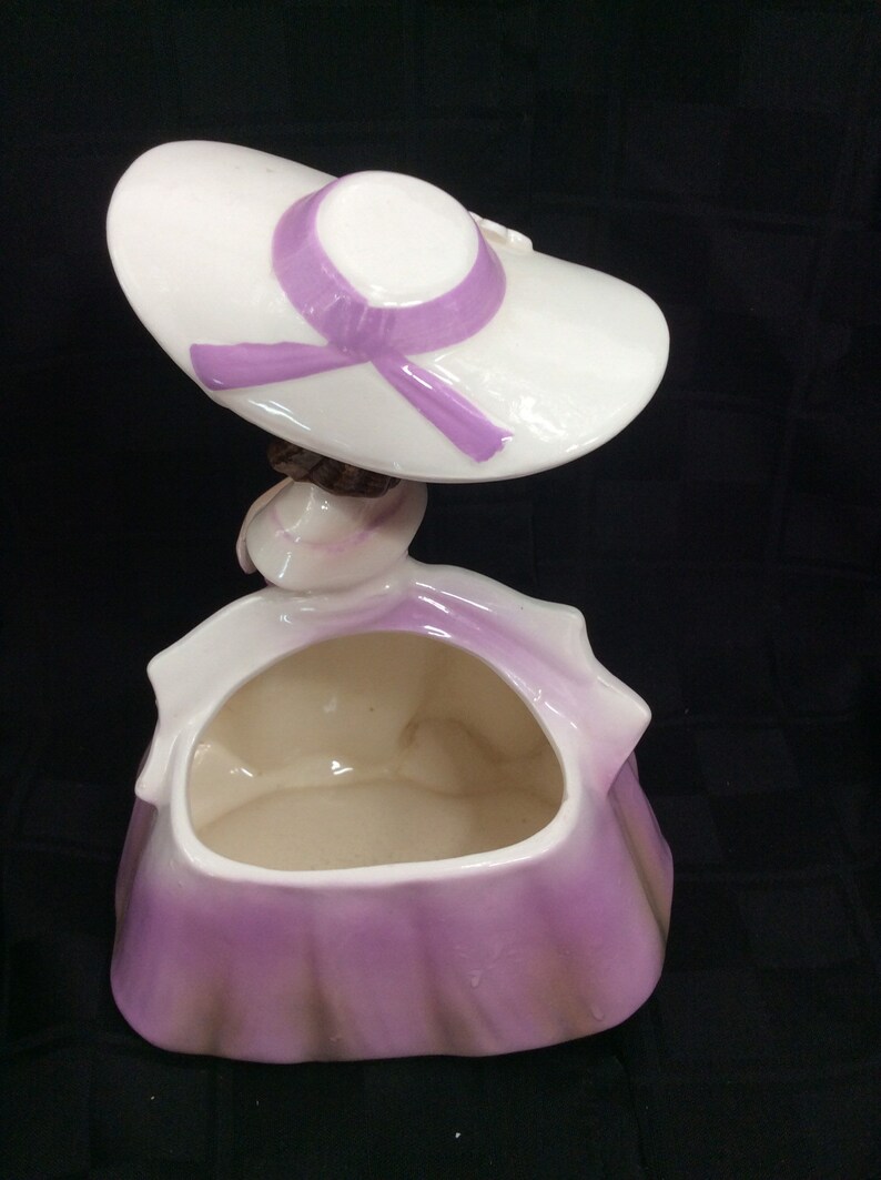 May include: A ceramic planter shaped like a woman wearing a white hat with a purple ribbon. The planter has a purple and white glaze and is designed to hold small plants or flowers.
