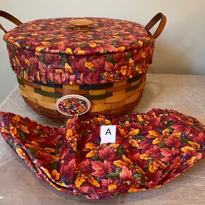 May include: A woven basket with a fabric lid and liner. The lid and liner feature a fall-themed print of red, orange, and yellow leaves with blue berries and acorns. The basket has a wooden handle and a wooden knob on the lid. The liner is folded and placed next to the basket. The letter "A" is written on a white tag.
