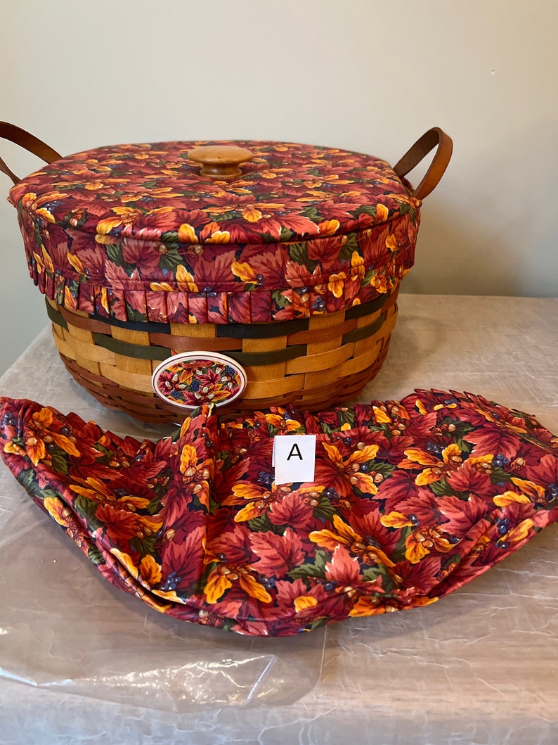 May include: A woven basket with a fabric lid and liner. The lid and liner feature a fall-themed print of red, orange, and yellow leaves with blue berries and acorns. The basket has a wooden handle and a wooden knob on the lid. The liner is folded and placed next to the basket. The letter "A" is written on a white tag.