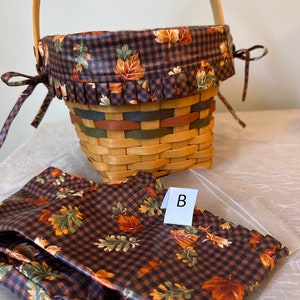 May include: A brown and black checked basket with a fall leaf print lining and a matching fabric liner. The basket has a wooden handle and a brown ribbon tied around the top. The liner has the letter "B" written on it.