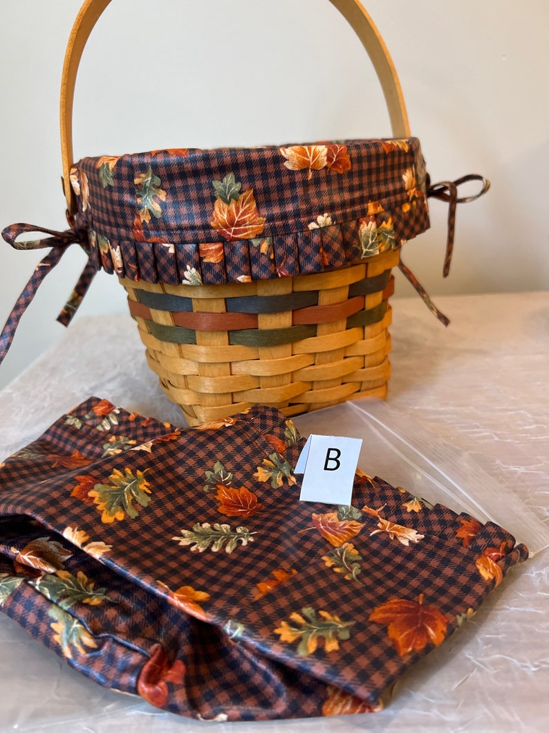 May include: A brown and black checked basket with a fall leaf print lining and a matching fabric liner. The basket has a wooden handle and a brown ribbon tied around the top. The liner has the letter "B" written on it.