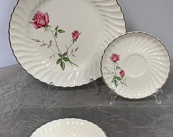 May include: A set of three off-white ceramic dishes featuring a swirling ribbed texture and a silver-toned rim. Each piece is decorated with a botanical illustration of pink roses with green leaves and stems, displayed on a large plate, a saucer, and a bowl.