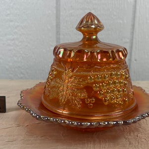 May include: A vintage orange glass butter dish with a grape design. The dish has a lid and a base.