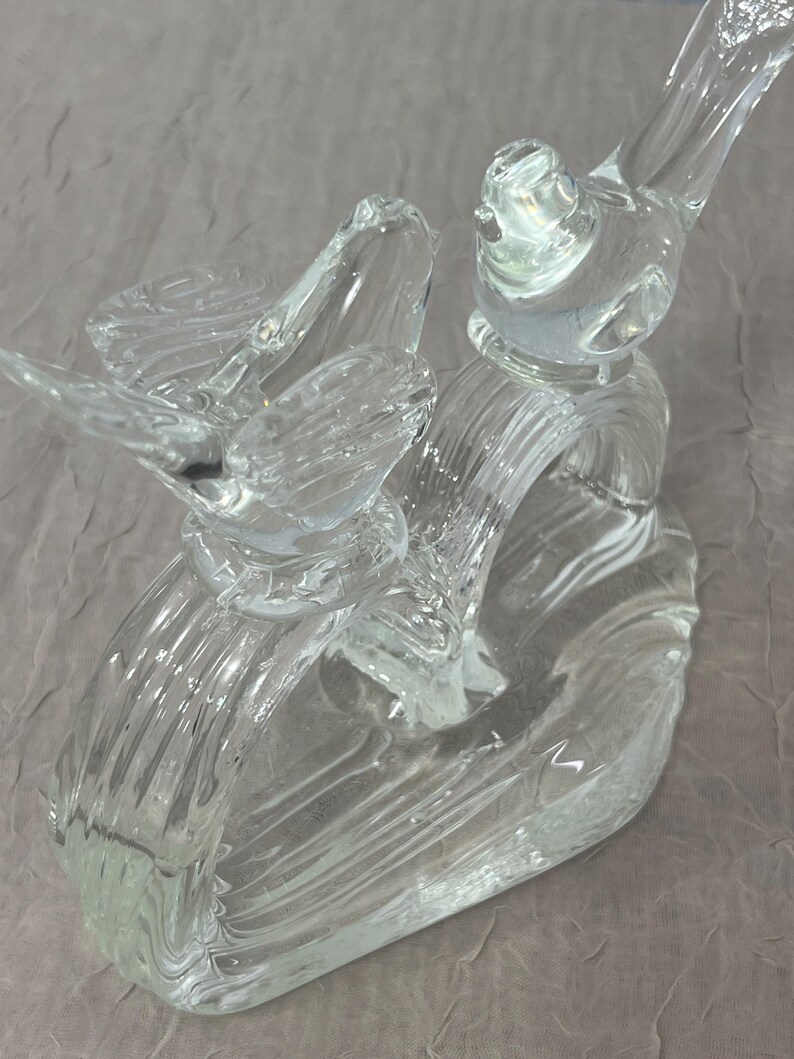 K&ouml;nnte beinhalten: Eine transparente Glasskulptur mit zwei V&ouml;geln, die auf einer gebogenen, gerippten Basis sitzen. Die V&ouml;gel haben detaillierte Fl&uuml;gel und Schn&auml;bel, und die Skulptur ist transparent. Es handelt sich wahrscheinlich um einen dekorativen Gegenstand.