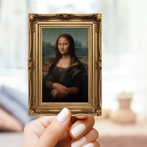 May include: A small, framed print of the Mona Lisa painting. The frame is ornate, gold-colored, and rectangular. The painting depicts a woman with a subtle smile, set against a landscape background. The frame is held by a person.