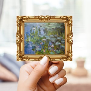 May include: A small, rectangular framed print featuring a water lilies painting in shades of blue, green, and pink. The ornate, gold-colored frame has detailed scrollwork. The artwork is held by a person with white nail polish.