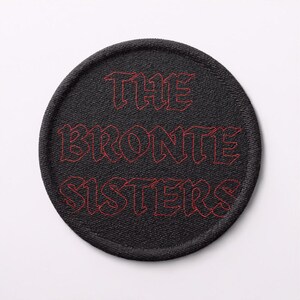 Puede incluir: Un parche redondo negro con texto bordado en rojo que dice "THE BRONTE SISTERS". El parche tiene una apariencia texturizada, con el texto en una fuente estilizada. El parche es probablemente para ropa o accesorios.