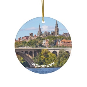 May include: A round ceramic ornament featuring a scenic view of Georgetown, Washington D.C. The ornament depicts a bridge over a river with buildings and trees in the background. The text "GEORGETOWN" is printed on the ornament.
