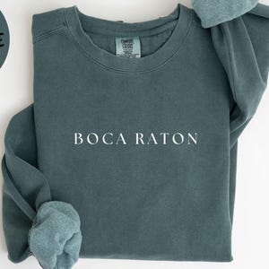 May include: A blue spruce colored sweatshirt with the text "BOCA RATON" in white. The words "BLUE SPRUCE" are in a circle in the upper left corner. The words "COMFORT COLORS 1566" are at the bottom.