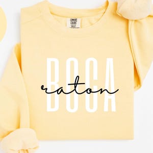 May include: A butter-yellow sweatshirt with the text "BOCA RATON" in white block letters and script. The words "BUTTER" and "COMFORT COLORS 1566" are also visible. The sweatshirt has long sleeves and a crew neck.