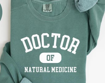 May include: Light green sweatshirt with the text "DOCTOR OF NATURAL MEDICINE" in white block letters. The sweatshirt is a Comfort Colors brand. The words "COMFORT COLORS 1566" are printed at the bottom.