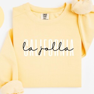 May include: A light yellow sweatshirt with the words "CALIFORNIA la jolla" printed on the front in white and black. The sweatshirt has long sleeves and a crew neck. The text "BUTTER" is in a yellow circle. The text "COMFORT COLORS 1566" is at the bottom.