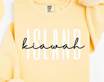 May include: A butter-yellow sweatshirt with the word "ISLAND kiawah ISLAND" in white block letters and cursive. The words "BUTTER" and "COMFORT COLORS 1566" are also visible in the image.