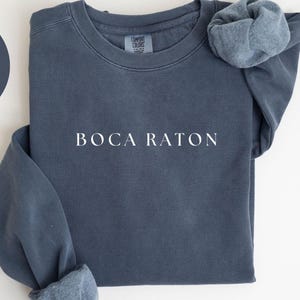 May include: Denim-colored sweatshirt with the text "BOCA RATON" in white. The sweatshirt has a crew neck and long sleeves with cuffed ends. The words "COMFORT COLORS 1566" are printed at the bottom. A denim-colored circle with the word "DENIM" is in the upper left.