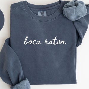 May include: A denim blue sweatshirt with the words "boca raton" in white script. The sweatshirt has a crew neck and long sleeves. A denim blue scrunchie rests on the shoulder. The words "COMFORT COLORS 1566" are printed at the bottom.