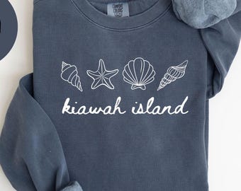 May include: A denim blue sweatshirt with white seashell graphics and the words "kiawah island". The sweatshirt has rolled-up sleeves and the words "COMFORT COLORS 1566" at the bottom. A denim circle with the word "DENIM" is in the upper left.