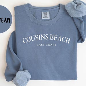 May include: A blue jean colored sweatshirt with the words "COUSINS BEACH EAST COAST" in white lettering. The sweatshirt has a crew neck and long sleeves. The words "BLUE JEAN" are in a circle on the left side of the image. The words "COMFORT COLORS 1566" are at the bottom.
