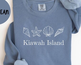 May include: A blue jean colored sweatshirt with white seashell graphics and the words "Kiawah Island". The text "Comfort Colors 1566" is printed at the bottom. A blue circle with the words "Blue Jean" is in the upper left.