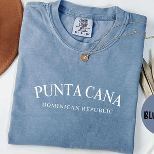 May include: A blue jean colored t-shirt with the words "PUNTA CANA DOMINICAN REPUBLIC" printed in white. The shirt is a Comfort Colors brand. A gold necklace with a pendant rests on the shirt. A brown hat and dried plants are in the background.