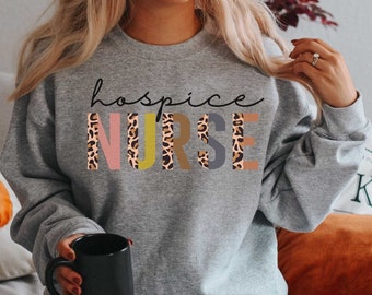 May include: A grey sweatshirt with the text "hospice NURSE" in colourful letters with a leopard print pattern.