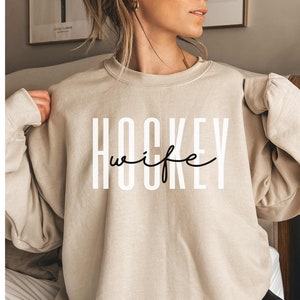 Op de afbeelding: Een beige sweatshirt met de tekst "HOCKEY wife" in zwart-witte letters.