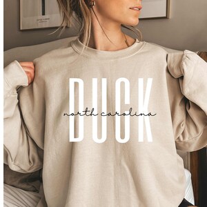 以下が含まれることがあります： ベージュのトレーナーに「DUCK north carolina」という白い文字が書かれています。トレーナーは人が着用しています。
