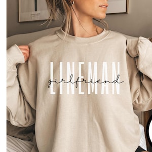May include: A beige sweatshirt with the text "LINEMAN girlfriend" printed in black and white. The sweatshirt is being worn by a person. The text "more colors available" is printed in a circle on a white background.