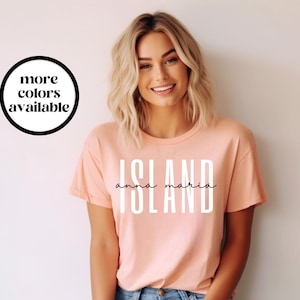 May include: A peach-colored t-shirt with the word "ISLAND" printed in white letters. The words "anna maria" are written in black script below the word "ISLAND".
