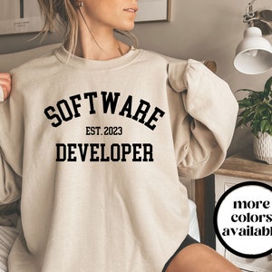 May include: A beige sweatshirt with the text "SOFTWARE DEVELOPER EST. 2023" in black, collegiate-style lettering. The sweatshirt has a crew neck and long sleeves. A circular graphic indicates more colors are available.