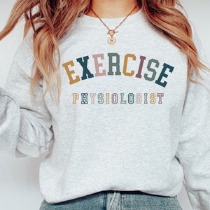 May include: A pale grey sweatshirt featuring the text "EXERCISE PHYSIOLOGIST" in a multi-coloured, collegiate-style font. The text uses shades of gold, pink, teal, and navy blue. The sweatshirt has long sleeves and a crew neck.