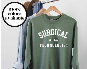 May include: A green sweatshirt with the words "SURGICAL TECHNOLOGIST EST. 2023" in white block letters. The sweatshirt is hanging on a wooden hanger. A circular graphic in the upper left corner reads "more colors available".