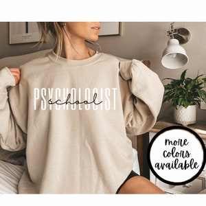 May include: A beige sweatshirt with the text "PSYCHOLOGIST school" printed in black on the front. The sweatshirt is being modeled by a person. The text "more colors available" is printed in black on a white circle in the lower right corner of the image.