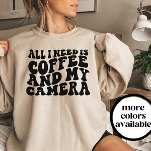May include: A beige sweatshirt with the text "All I need is coffee and my camera" in black retro-style lettering.