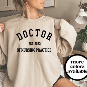 May include: A beige sweatshirt with the text "DOCTOR EST. 2023 OF NURSING PRACTICE" printed on the front. The text is in a black, bold font.