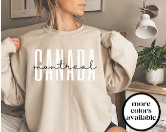 May include: A beige sweatshirt with the text "CANADA" in white letters and "montreal" in black script underneath. The text is centered on the sweatshirt. The sweatshirt is being worn by a person. The text "more colors available" is in a circle in the lower right corner of the image.