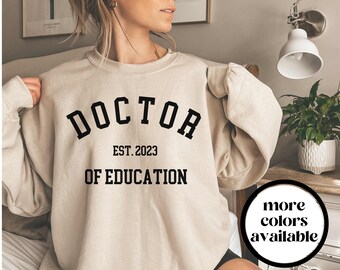 Puede incluir: Una sudadera beige con el texto "DOCTOR EST. 2023 OF EDUCATION" impreso en negro.