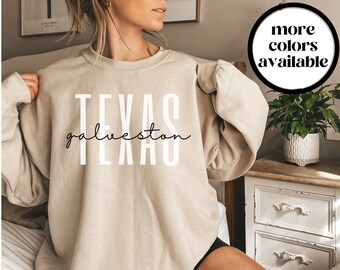 May include: A beige sweatshirt with the words "TEXAS galveston TEXAS" in white block and script lettering. A black circle with the words "more colors available" is in the upper right corner. The sweatshirt is part of a clothing collection.