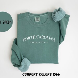 May include: A light green sweatshirt with the text "NORTH CAROLINA TARHEEL STATE" in white. The words "LIGHT GREEN" are in a circle above the sweatshirt. The words "COMFORT COLORS 1566" are below the sweatshirt.