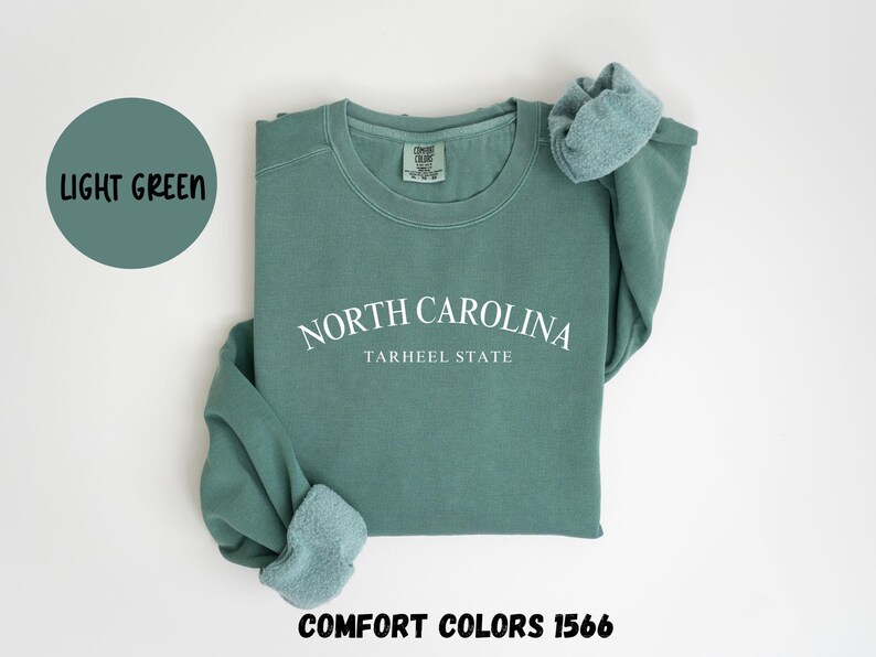 May include: A light green sweatshirt with the text "NORTH CAROLINA TARHEEL STATE" in white. The words "LIGHT GREEN" are in a circle above the sweatshirt. The words "COMFORT COLORS 1566" are below the sweatshirt.