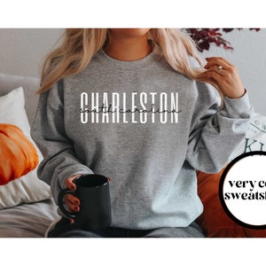 May include: A gray sweatshirt with the text "CHARLESTON south carolina" in white lettering. The sweatshirt is being worn by a person holding a black mug. The text "very cozy sweatshirts" is visible in a circle in the lower right corner of the image.