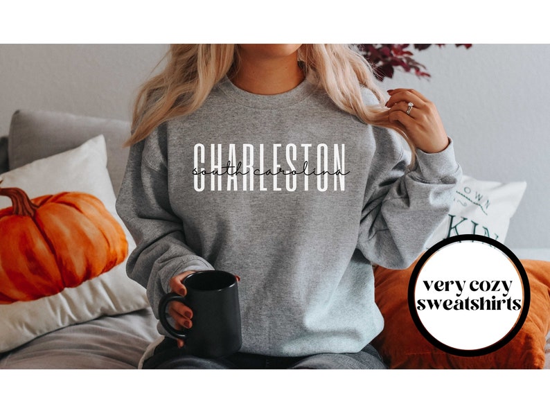 May include: A gray sweatshirt with the text "CHARLESTON south carolina" in white lettering. The sweatshirt is being worn by a person holding a black mug. The text "very cozy sweatshirts" is visible in a circle in the lower right corner of the image.