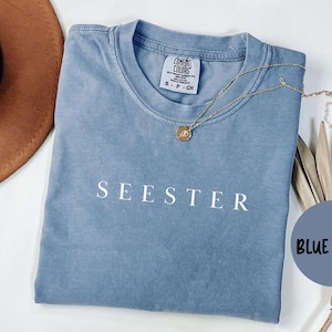 May include: A blue jean-colored t-shirt with the word "SEESTER" printed in white. The shirt is folded and has a Comfort Colors tag. A gold necklace with a pendant rests on the shirt. A brown hat is partially visible.